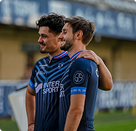 Deux joueurs se font une accolade et regardent hors champ, l'air confiant et souriants. L'action se déroule dans un stade